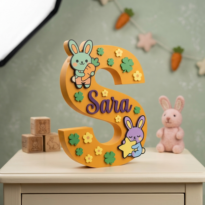 Lettera S decorativa da appoggio color giallo senape personalizzata con il nome Sara in viola, decorata in rilievo con teneri coniglietti, fiori e quadrifogli verdi, per arredamento cameretta bambina.