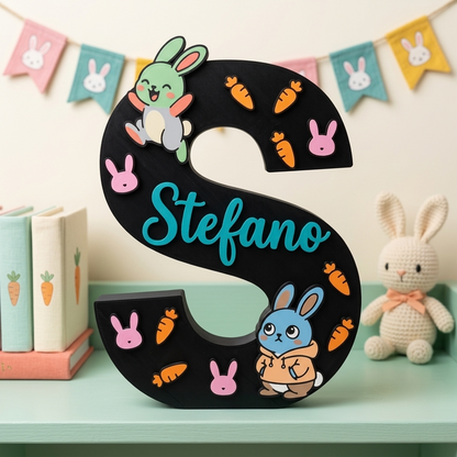 Lettera S decorativa nera da appoggio personalizzata con il nome Stefano in azzurro. Decorazione per cameretta a tema coniglietti, arricchita con figure di conigli colorati, faccine rosa e piccole carote arancioni in rilievo.