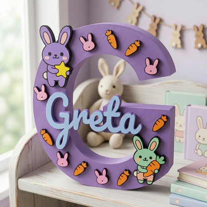 Lettera G lilla decorativa in 3D personalizzata con il nome Greta in azzurro. Decorazione per cameretta a tema coniglietti, applicati in rilievo insieme a carote e una stella gialla.