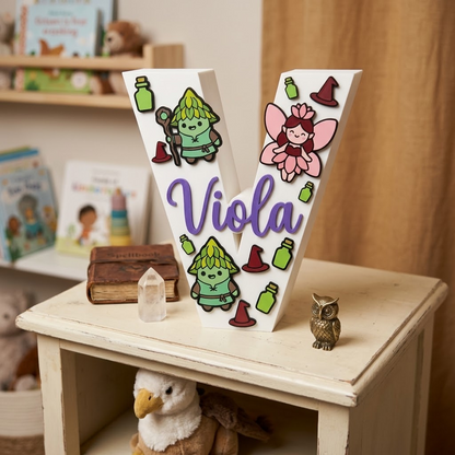 Lettera V decorativa 3D personalizzata con nome Viola in corsivo lilla. Iniziale bianca per cameretta bambina con decorazioni a tema magia: folletti del bosco, fata rosa, pozioni e cappelli da mago.