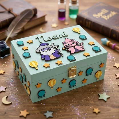 Scatola in legno azzurra personalizzata con nome Noemi in rilievo, decorata con applicazioni 3D a tema magia tra cui un mago, un gatto magico, stelle, mongolfiere e sfere di cristallo.
