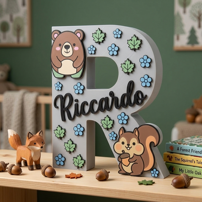 Lettera R decorativa grigia per cameretta, personalizzata con il nome Riccardo in corsivo nero. Design a tema animali del bosco con decorazioni in rilievo di un orsetto, uno scoiattolo con ghianda, foglie verdi e fiorellini azzurri.