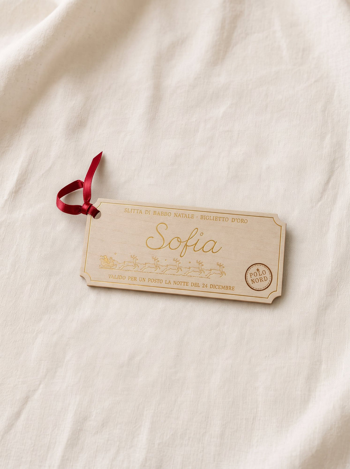 Biglietto d'oro con il nome Sofia su lino panna, nastro rosso nel foro e timbro Polo Nord inciso nel legno