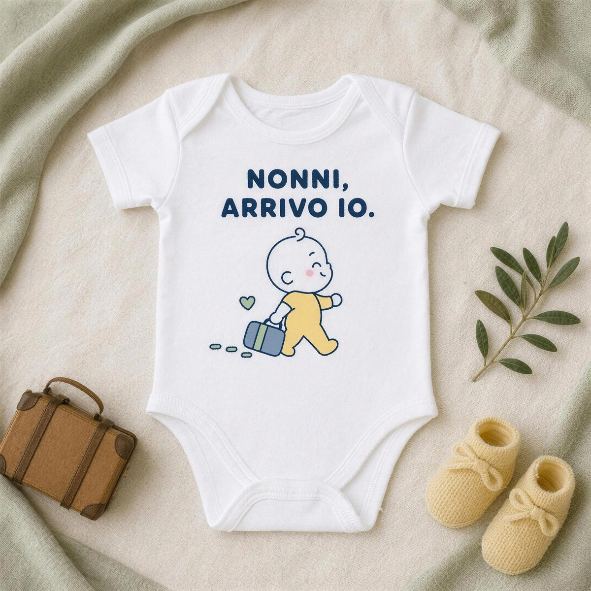 Body bianco con la frase «Nonni, arrivo io.» accanto a una valigetta in cuoio, scarpine gialle e un rametto d'ulivo