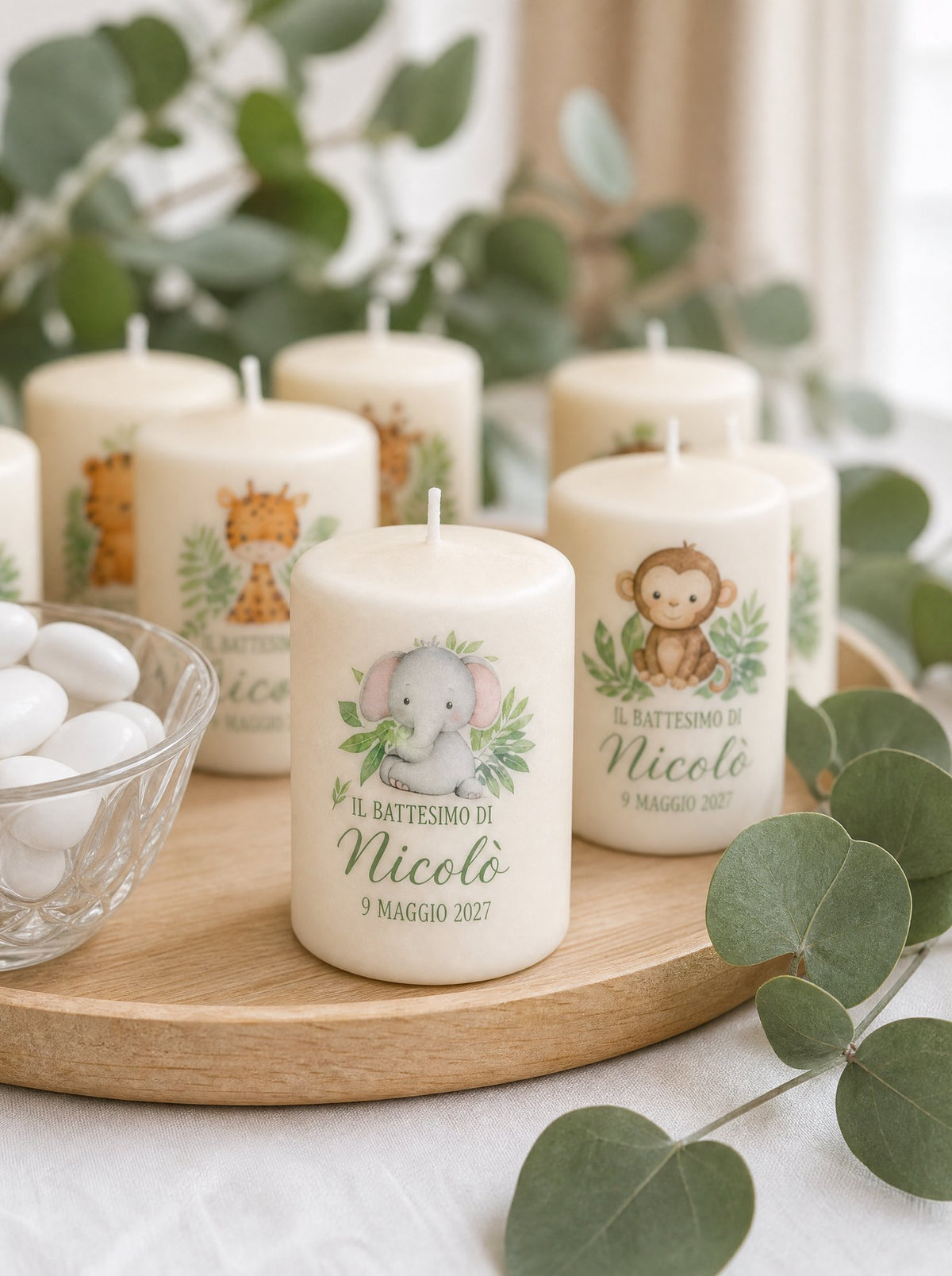 Candele battesimo su un vassoio di legno con rami di eucalipto e confetti, elefantino e scimmietta in primo piano