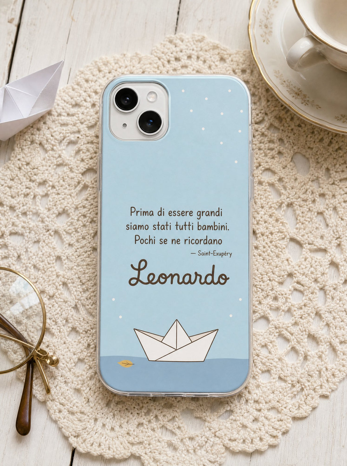 Cover per i nonni con una barchetta di carta, frase di Saint-Exupéry e nome Leonardo, su un iPhone 15 visto dall'alto