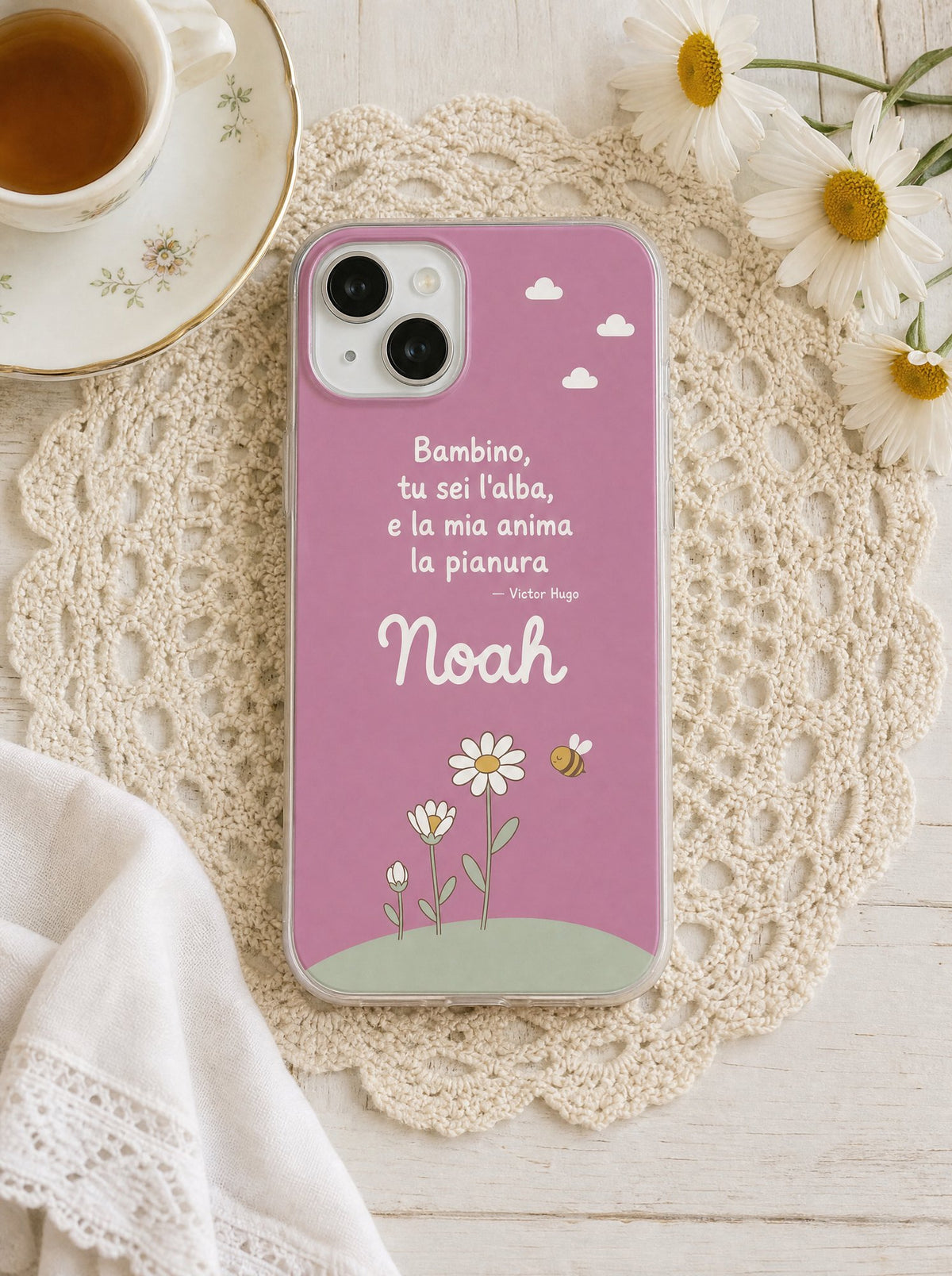 Cover per i nonni con margherite e un'ape, frase di Victor Hugo e nome Noah, su un iPhone 15 visto dall'alto